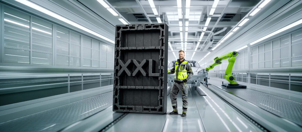 A worker in high-visibility industrial gear stands next to a large black XXL EPP load carrier inside a modern automated production hall, with a green robotic arm operating nearby.